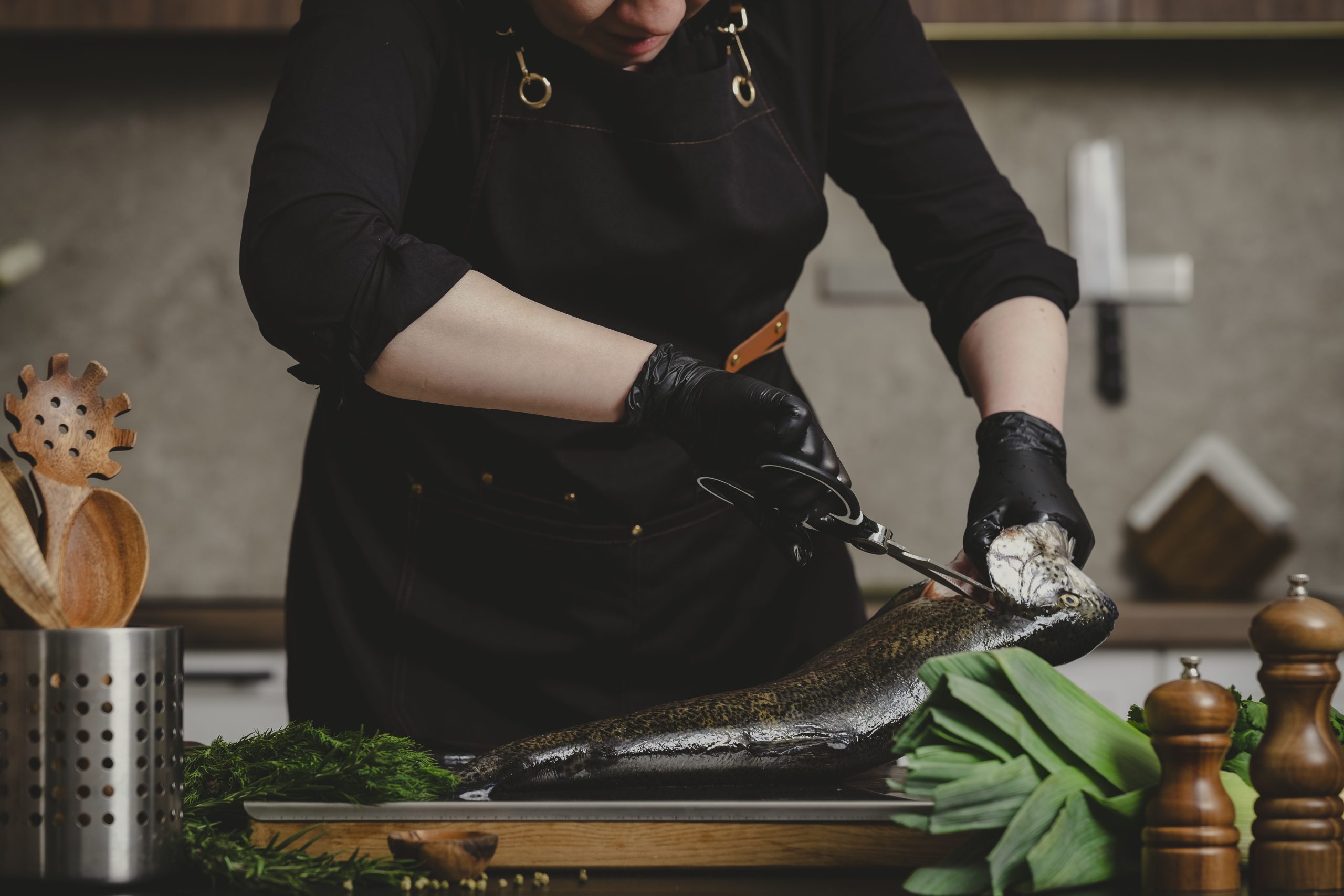 Hands of housewife cutting raw trout fish head with kitchen scissors. Precision gutting technique and seafood processing concept.